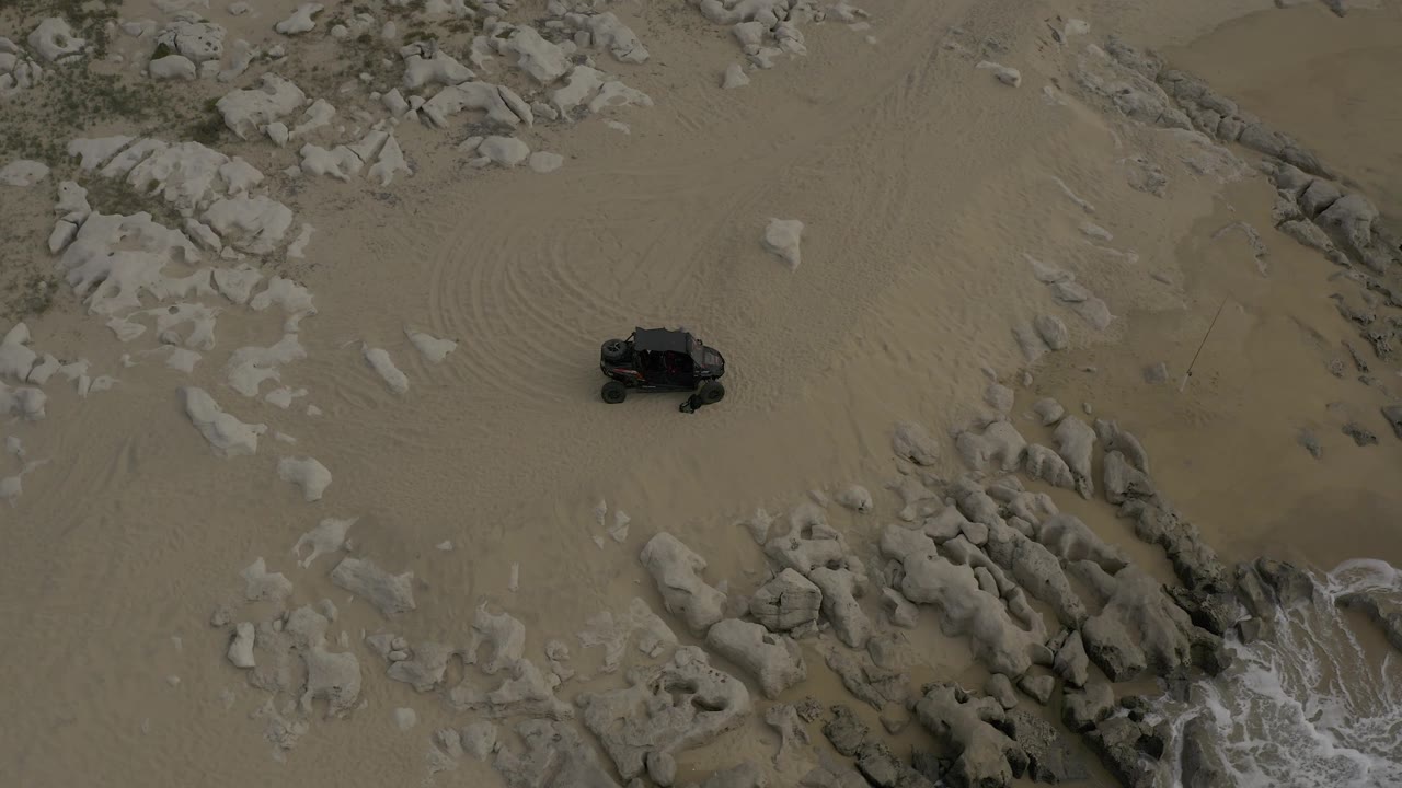 Top-down view from Baja California Sur, Mexico, showing a dark off-road vehicle on pale sand beside rock formations and white surf, with no visible people.