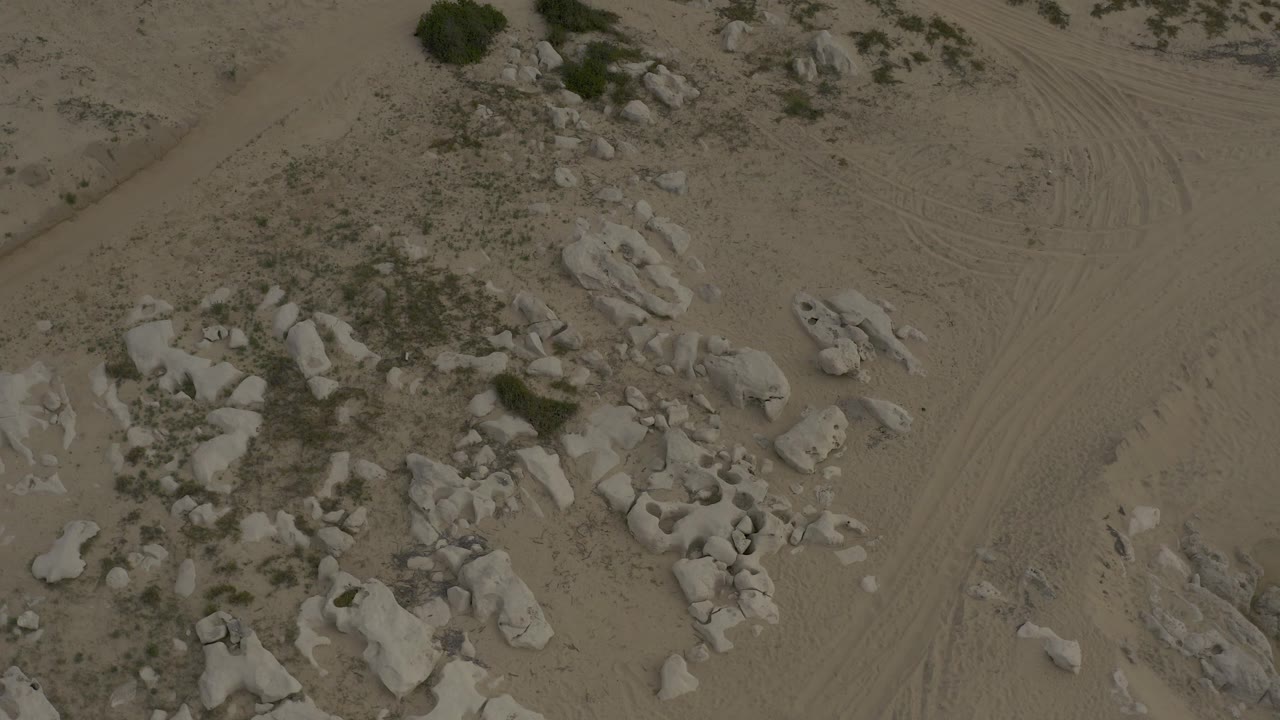 Low aerial frame over Baja California Sur desert terrain with pale rock clusters, scattered scrub, sandy tracks, and no visible people.
