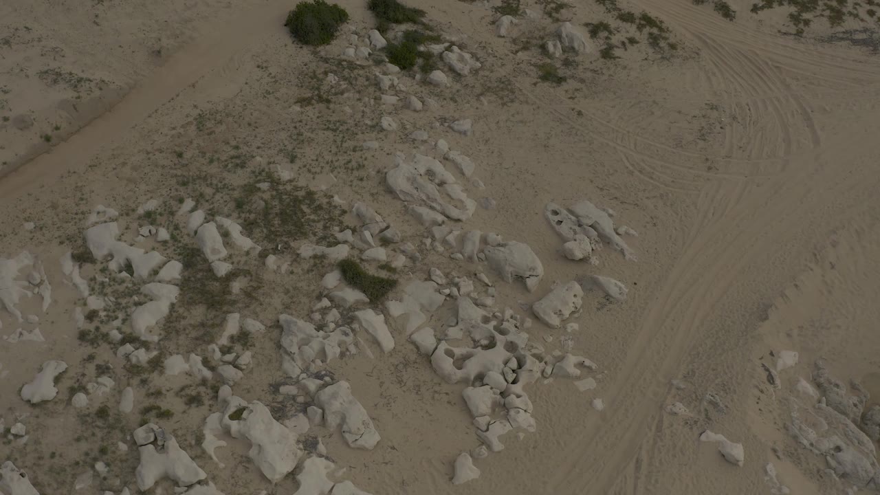 A later low aerial view in Baja California Sur shows a denser rock patch beside a pale dirt path, with sparse shrubs and no visible people.
