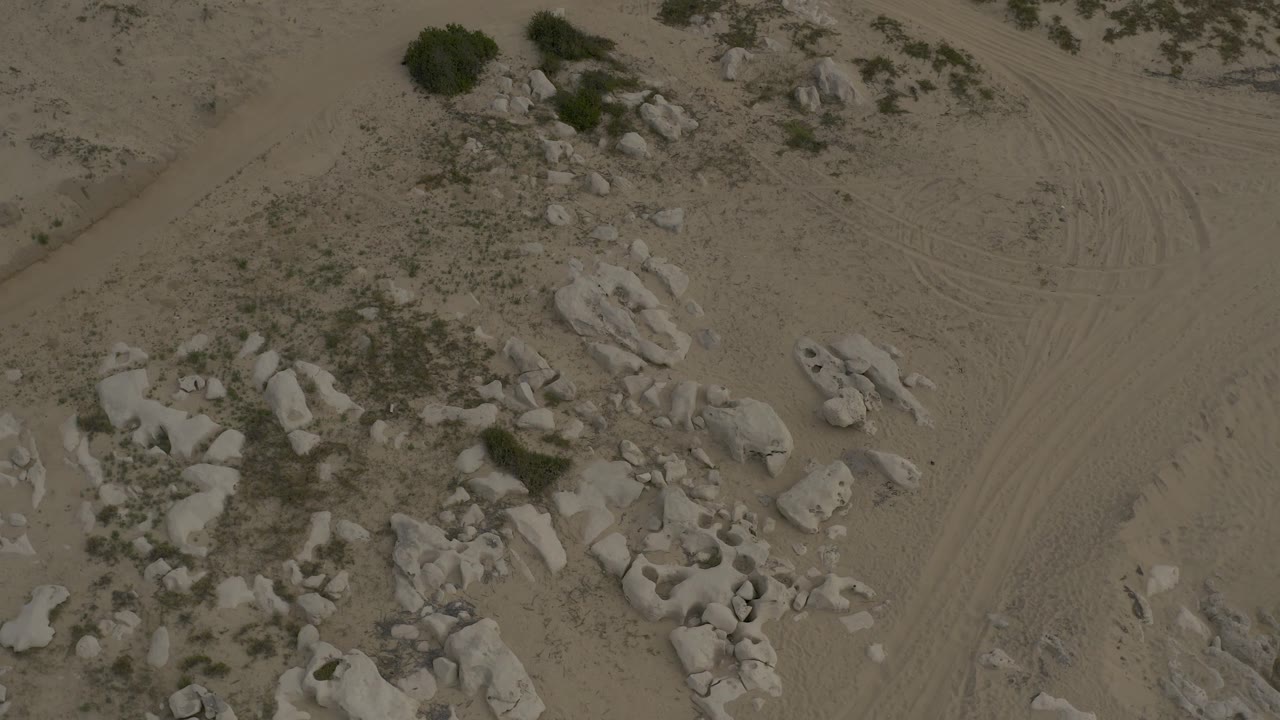 Low aerial view over Baja California Sur desert with rock outcrops on one side, curved tire marks in the sand, scattered scrub, and no visible people.