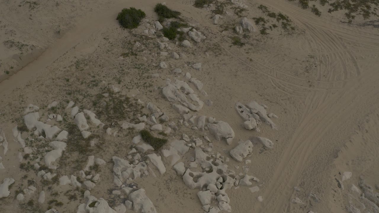 Low aerial frame in Baja California Sur with a diagonal dirt track cutting through sandy ground near boulders, scrub patches, and no visible people.