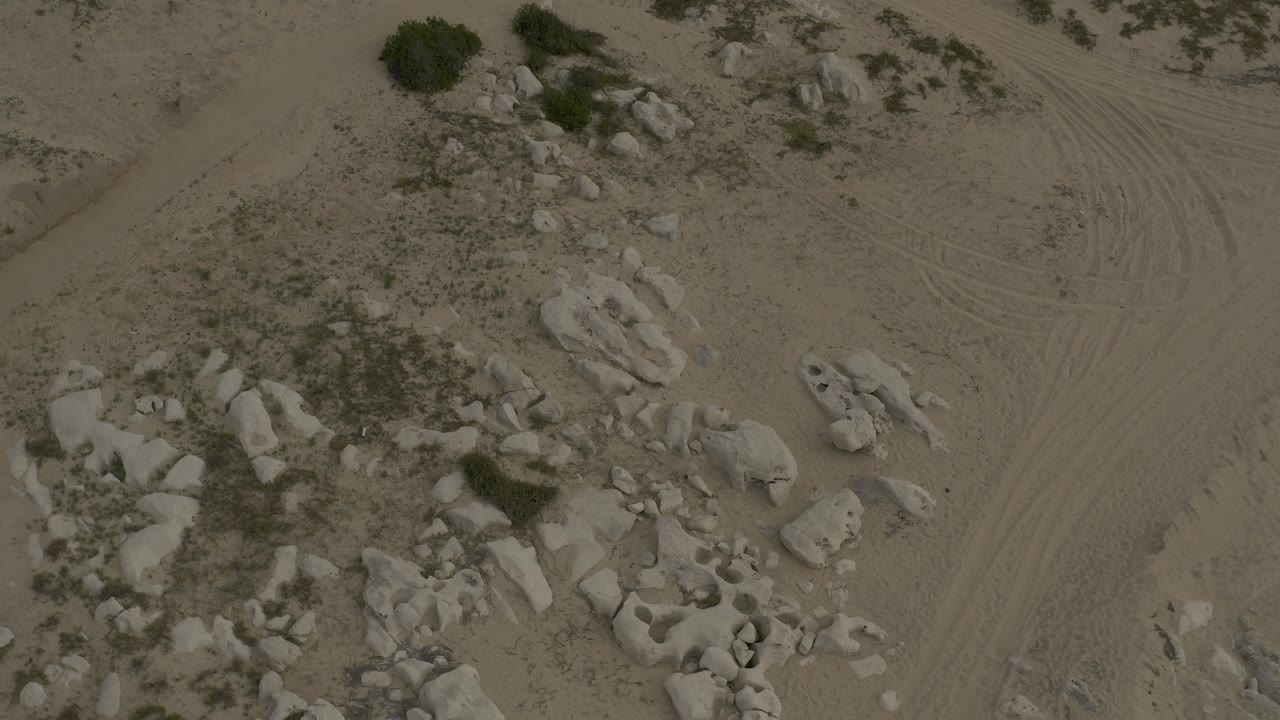 Overhead aerial frame of Baja California Sur desert sand with pale rock clusters, scattered shrubs, faint vehicle tracks, and no visible people.