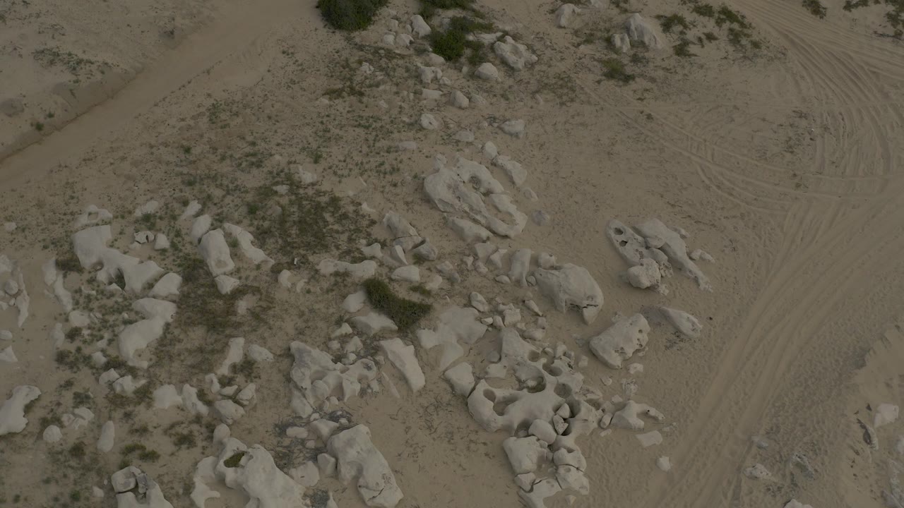 Opening overhead frame of rocky sandy terrain in Baja California Sur, Mexico, with pale boulder clusters, sparse scrub, tire marks, and no visible people.