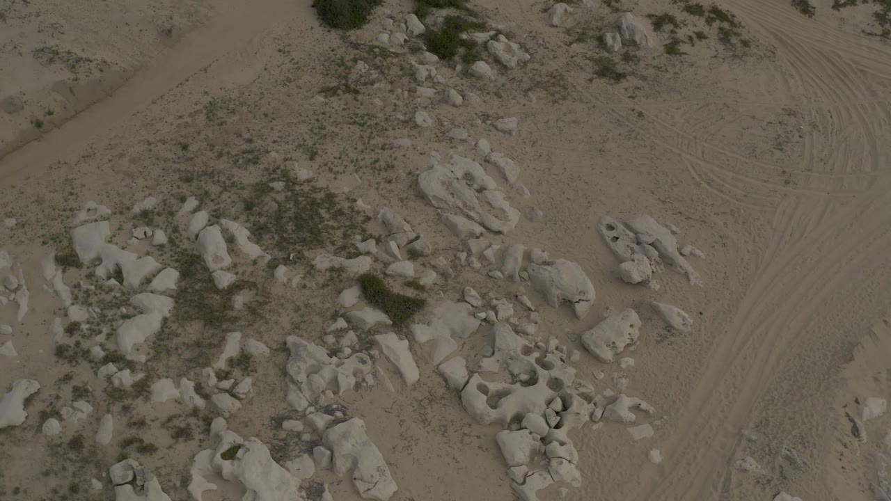 Second overhead frame in Baja California Sur, Mexico, showing a larger sandy lane with curved tire tracks beside scattered pale rocks and low scrub, with no visible people.