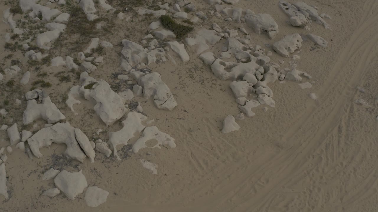 Overhead frame in Baja California Sur Mexico with pale rocks spread across tan sand, scrub patches near the rocks, visible track lines, and no visible people.