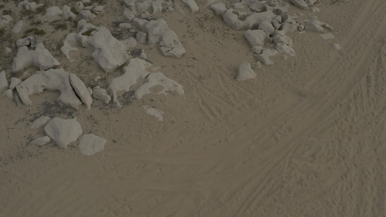 Downward-looking frame of sandy ground in Baja California Sur, Mexico, with pale boulders clustered near the upper left and tire marks crossing the sand, no visible people.
