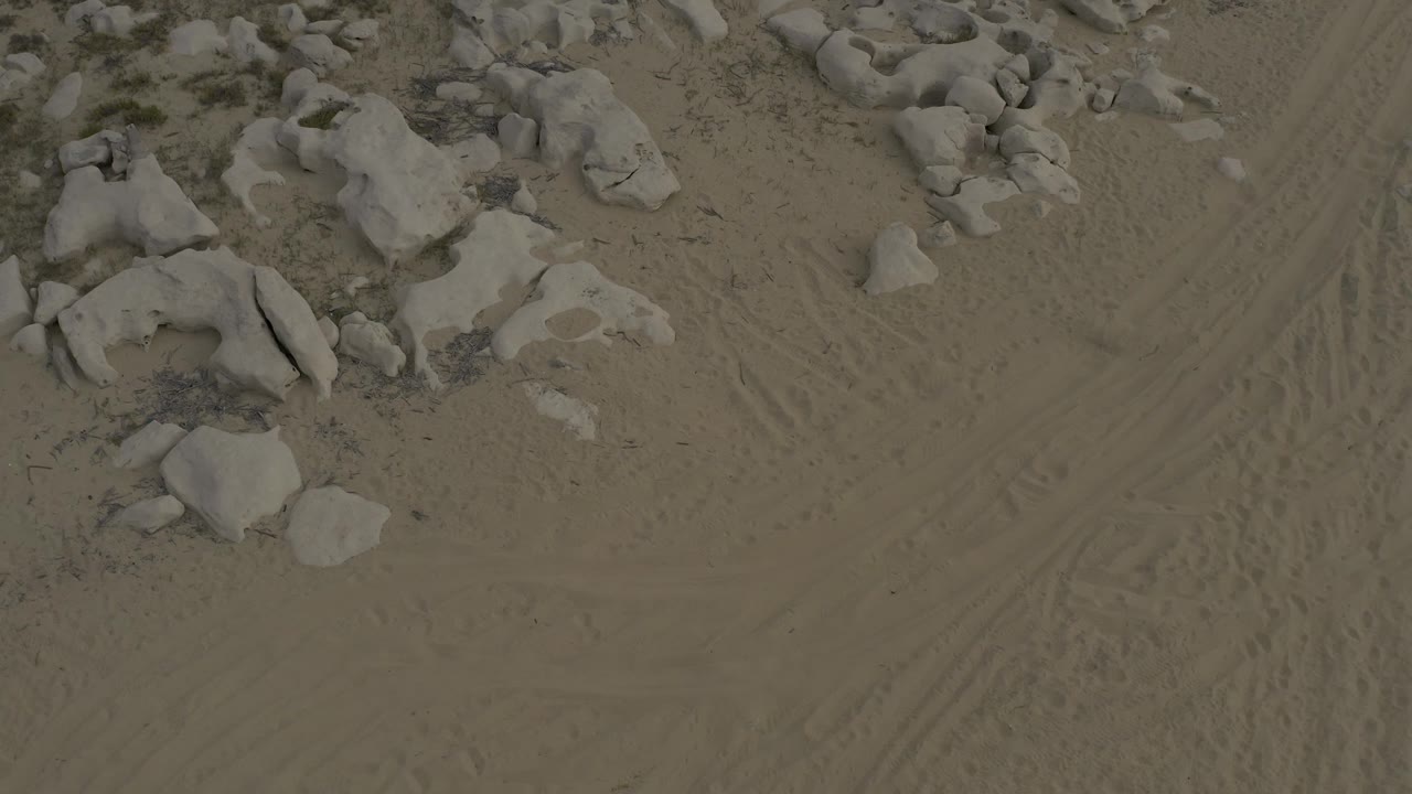 A high view over Baja California Sur sand shows rounded boulders along the upper left area and curved vehicle tracks running through the center, no visible people.