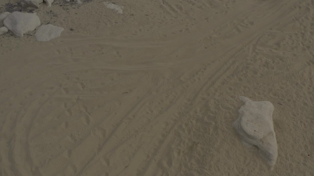 Overhead frame from Baja California Sur, Mexico showing tan sand, faint tire and foot tracks, several pale rocks near the upper edge, and no visible people.