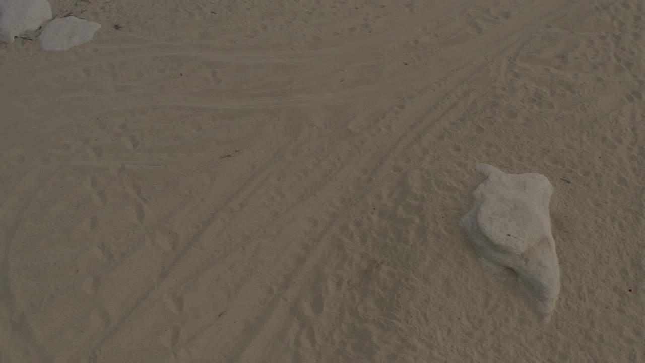 Aerial-looking frame over Baja California Sur sand with diagonal tire marks, scattered footprints, pale rocks at the top edge, and no visible people.