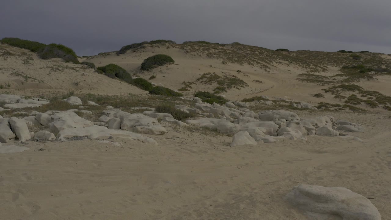 Wide view of a Baja California Sur beach with pale shoreline rocks, scrub-covered dunes, flat sand, and no visible people.