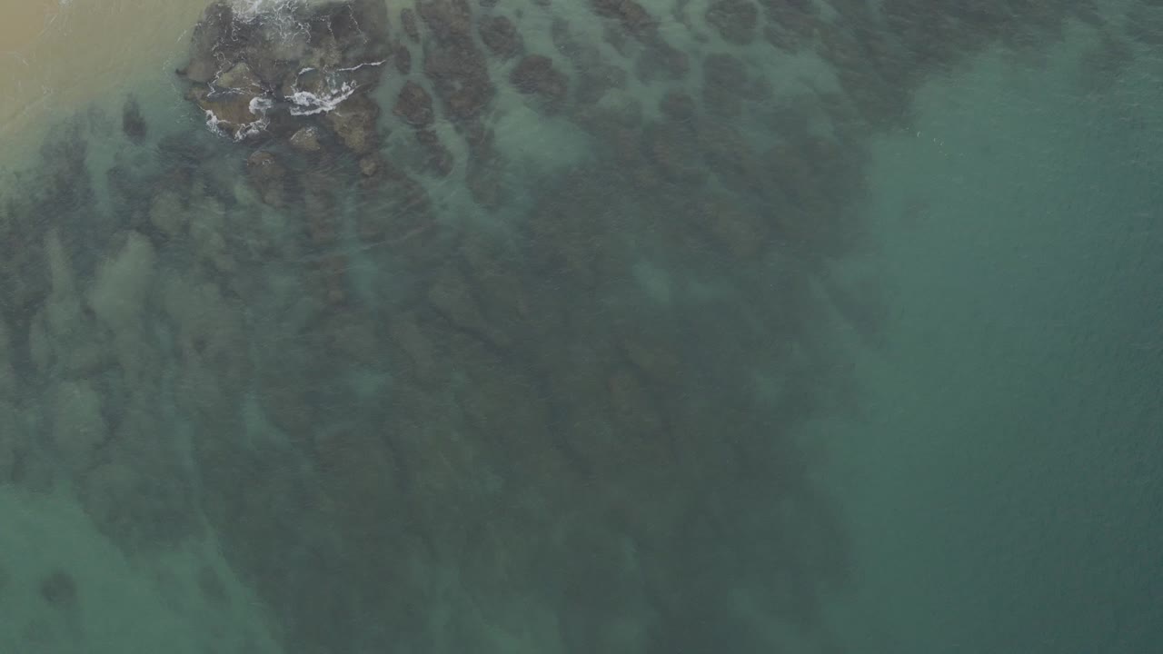 Overhead teal ocean with dark submerged rocks and a sandy corner