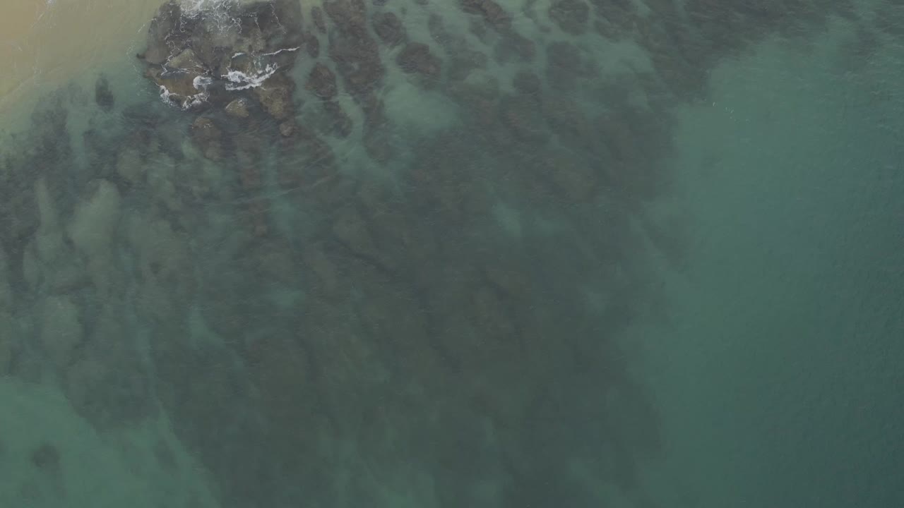 Aerial frame over Baja California Sur water showing dark reef-like shapes below the surface, a pale sand strip at upper left, and no visible people.