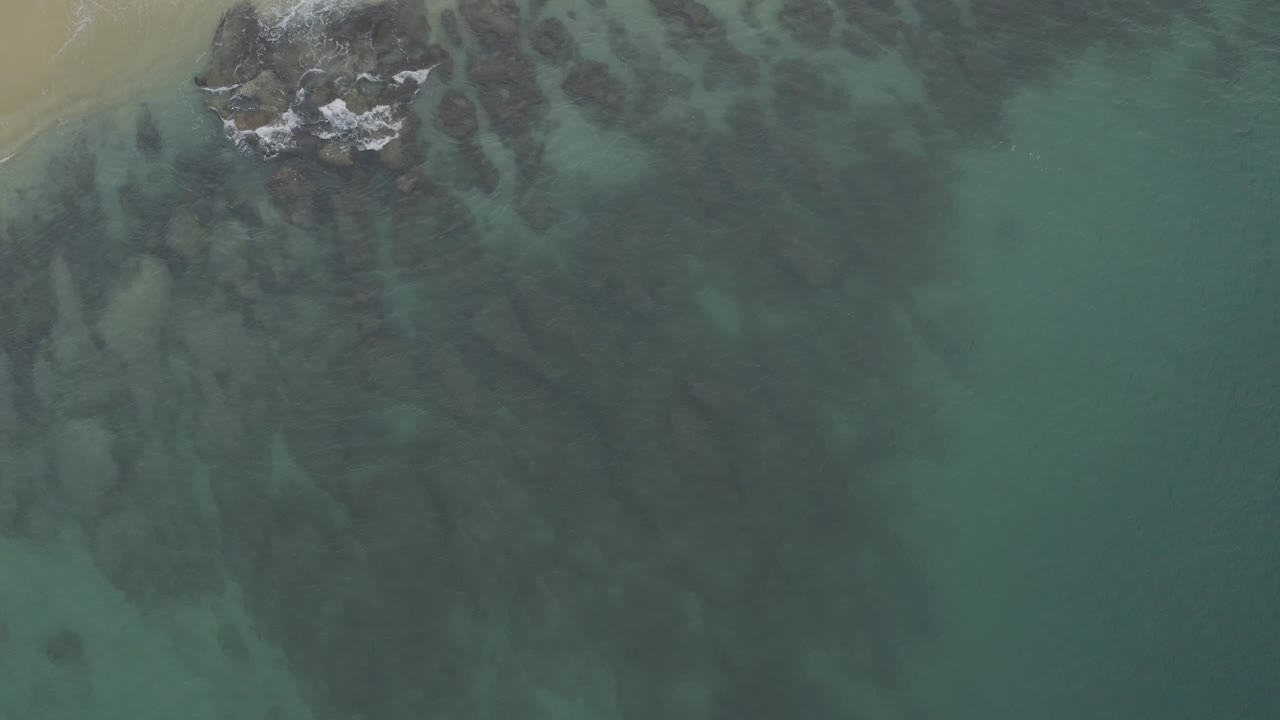 Top-down Baja California Sur shoreline view with a rocky area near the upper left, faint white surf, dark underwater streaks, and no visible people.