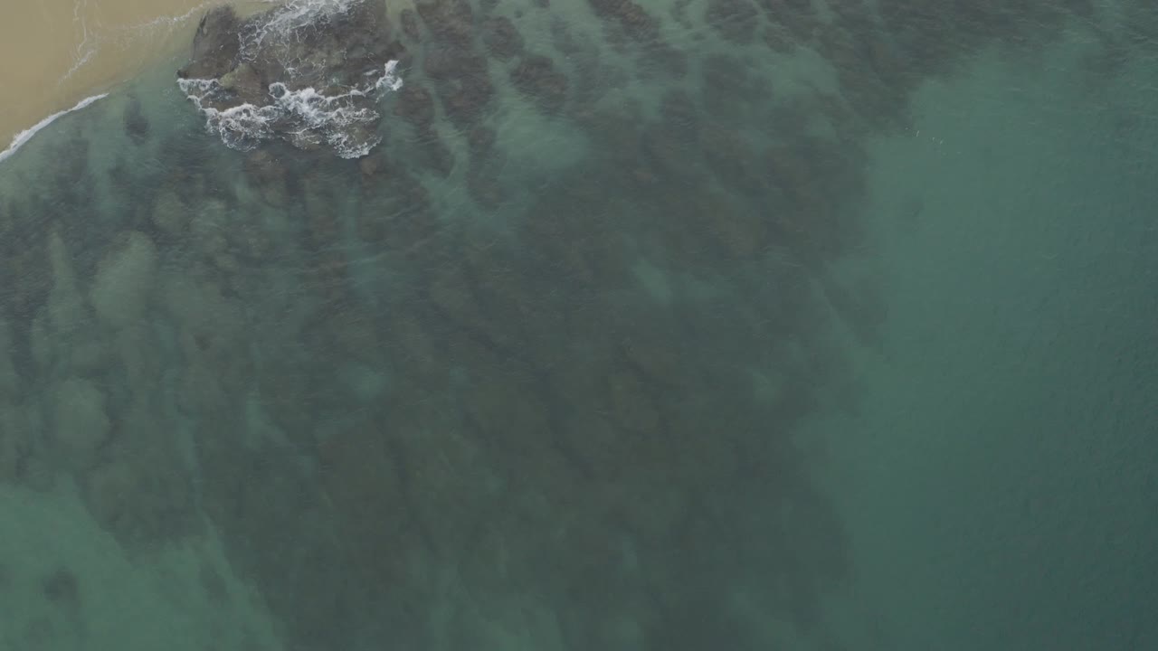 A Baja California Sur aerial still where a white incoming wave reaches the exposed rocks near the sand, with green water and no visible people.