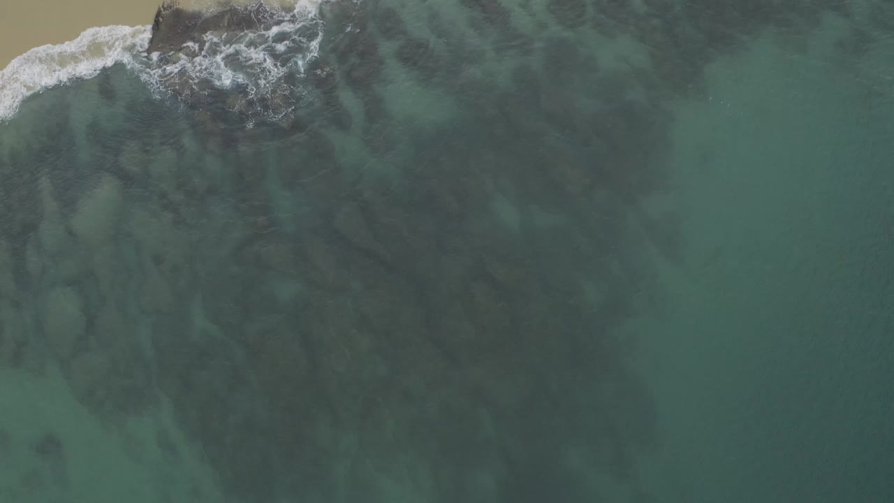 Aerial frame over Baja California Sur coastal water where dark submerged rocks fill the center and a small line of white foam reaches in from the upper left, no visible people.