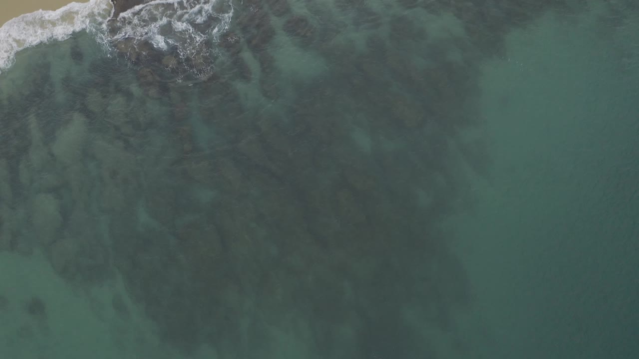 Top-down view of Baja California Sur shoreline water with foamy rocks across the upper left and a wide field of blue-green ocean extending to the right, no visible people.