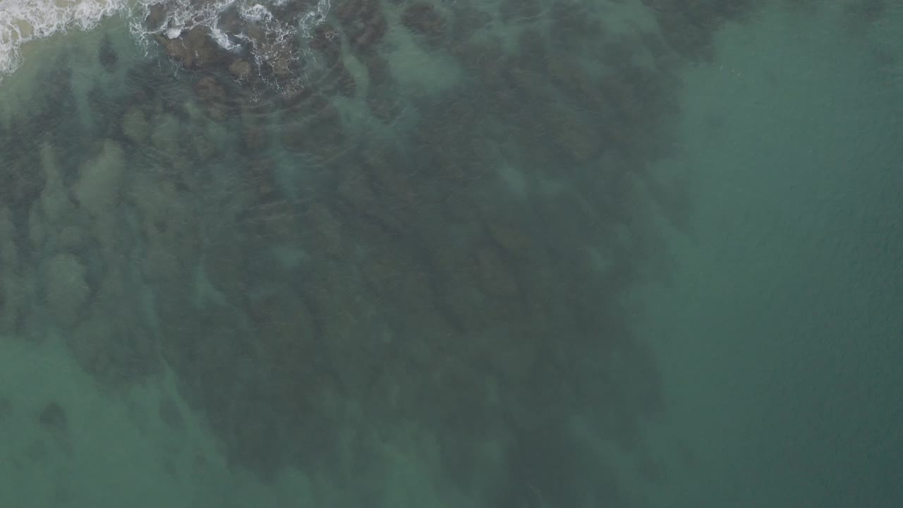 Clear shallow sea in Baja California Sur seen from above, with darker underwater rock forms drifting through the center and faint foam tucked into the upper left corner, no visible people.