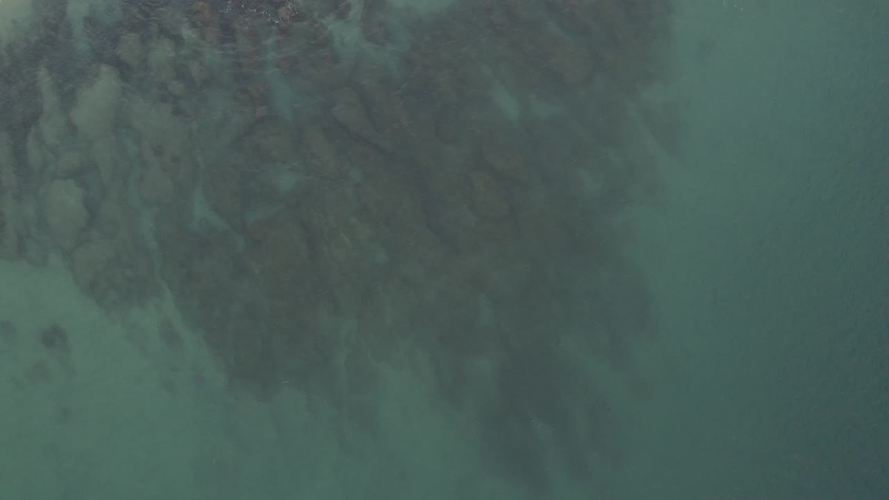 Top-down aerial frame of shallow blue-green water and muted reef shapes along the upper right in Baja California Sur, Mexico, with no visible people.