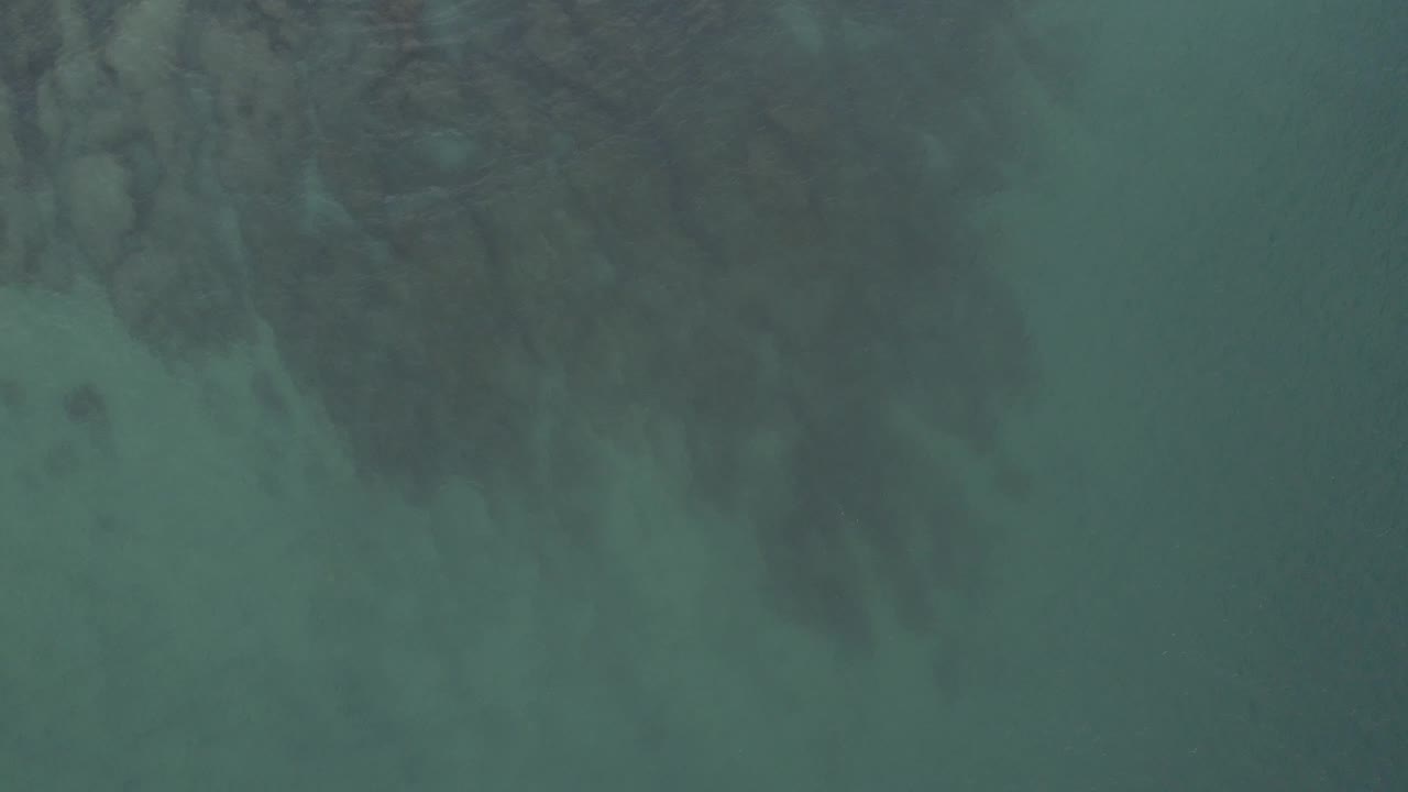 A high, downward view over Baja California Sur ocean water shows clustered dark reef forms under a green surface and no visible people.