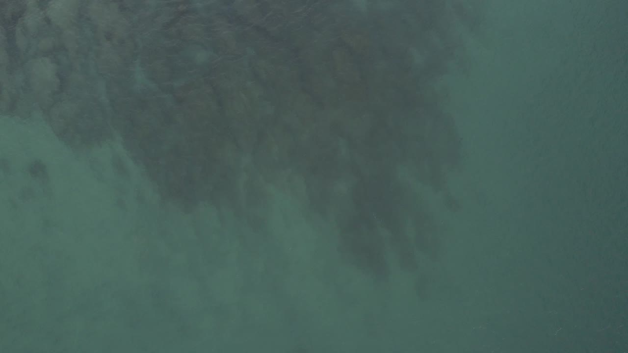 Muted green ocean water in Baja California Sur, Mexico, shows reef shadows gathered along the upper frame while the lower area remains more open, with no visible people.