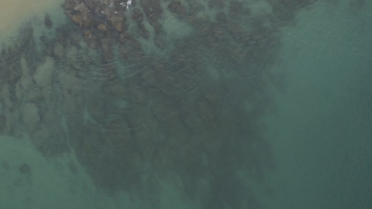 Overhead blue-green water with dark submerged rock patterns near a sandy shore.