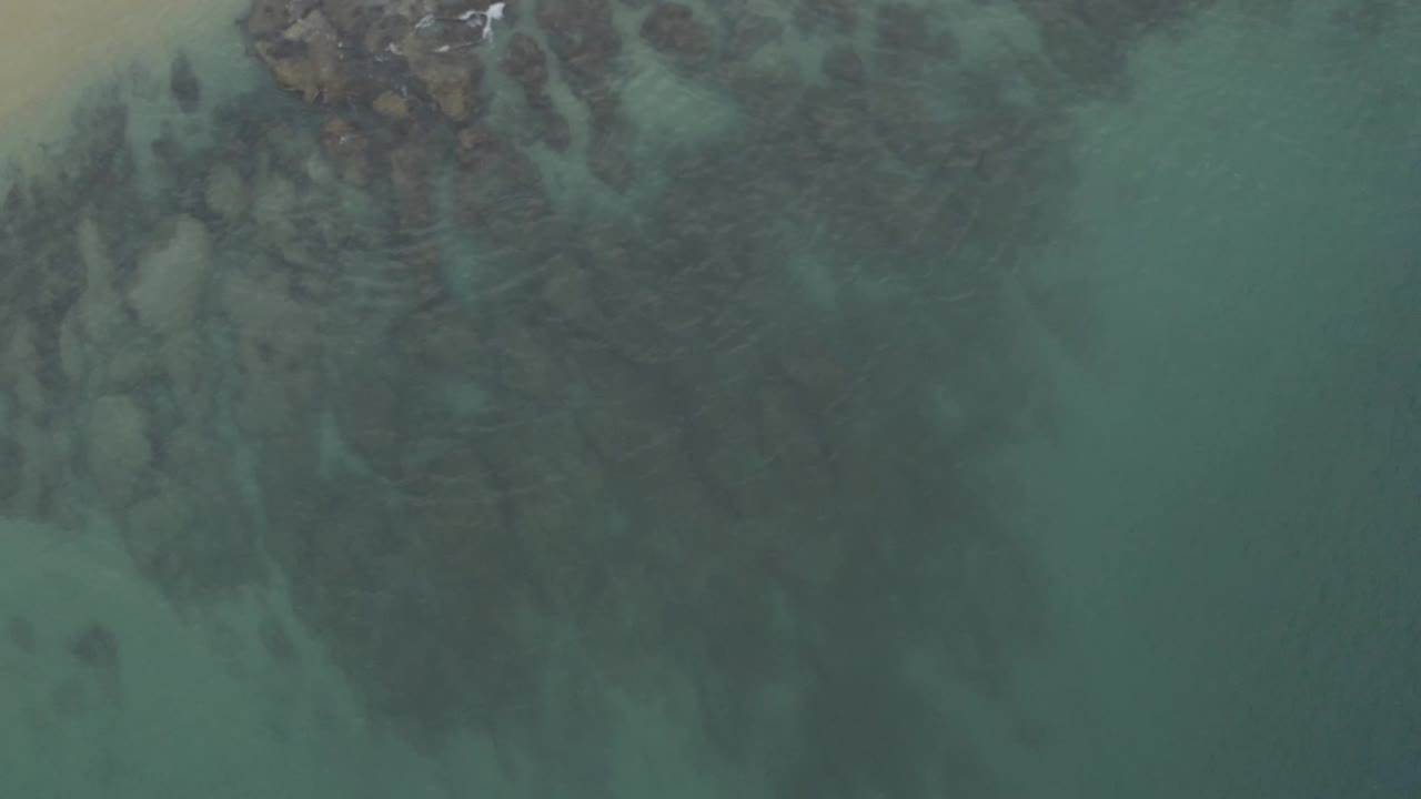 Aerial frame from Baja California Sur, Mexico with shallow blue-green water, dark underwater formations, a small sandy edge, and no visible people.