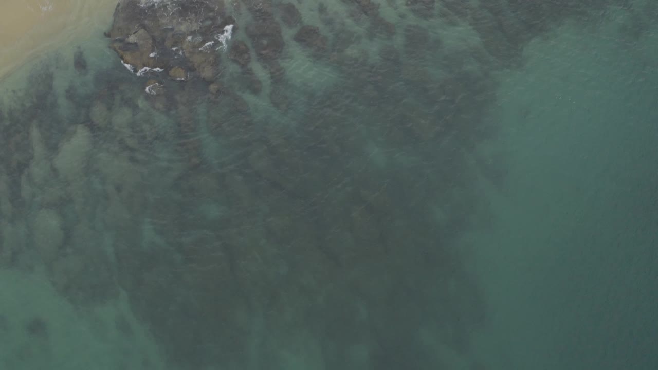 Top-down view in Baja California Sur, Mexico showing tan beach at the upper left, grey rocks at the surf line, blue-green water, and no visible people.