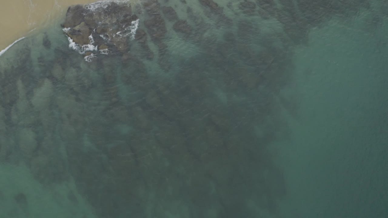 Overhead Baja California Sur shoreline frame with a tan beach corner, grey rocks, white wave foam, blue-green water, and no visible people.