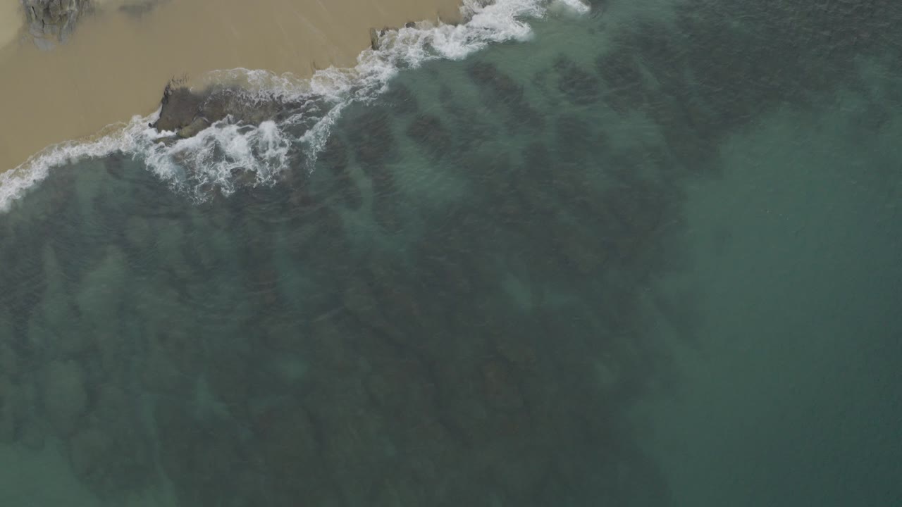Aerial view over Baja California Sur, Mexico, with blue-green water below a tan beach, dark submerged rock patches, white surf at the shore, and no visible people.