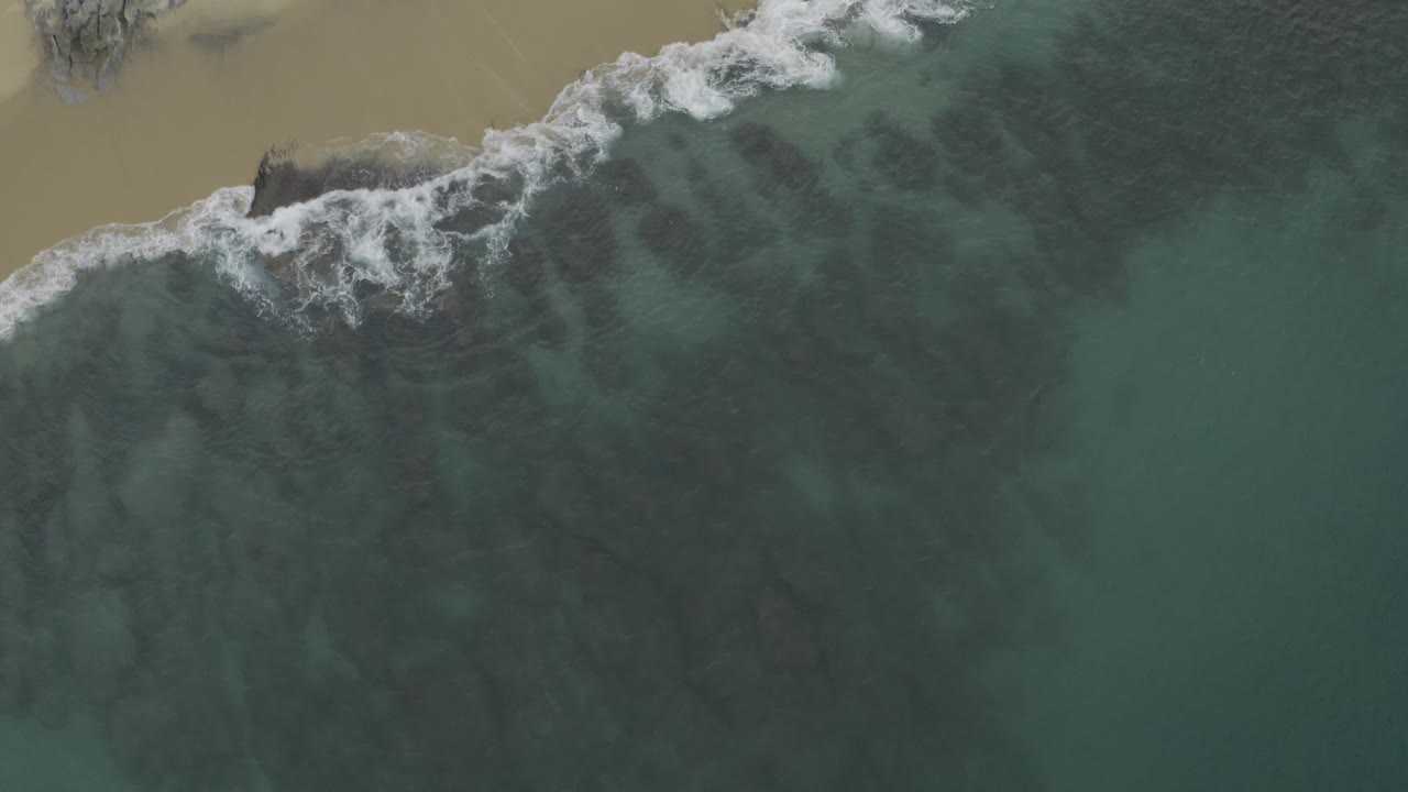 Drone view over Baja California Sur, Mexico, with white foam thickening near a rocky patch on the sandy shore and open blue-green ocean filling the lower frame, with no visible people.
