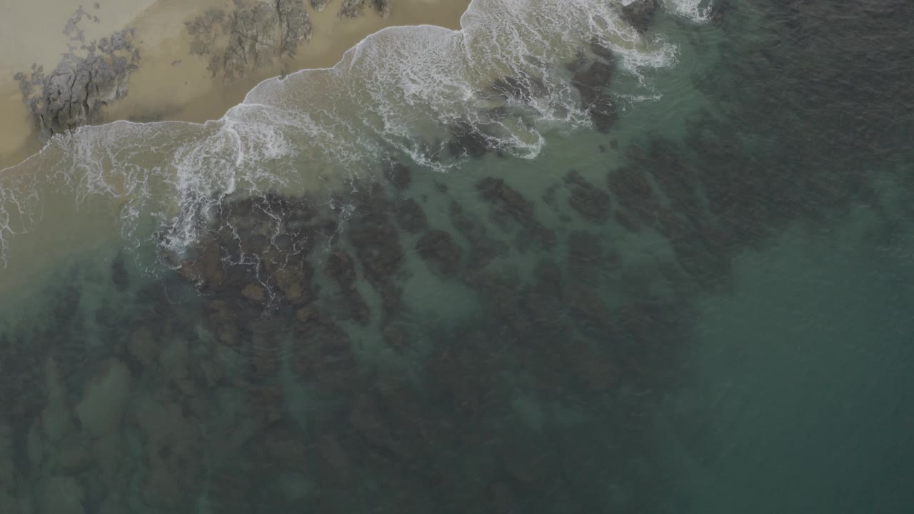Top-down view of Baja California Sur coast where a white wave curls along the sand and darker water fills the right side, with no visible people.