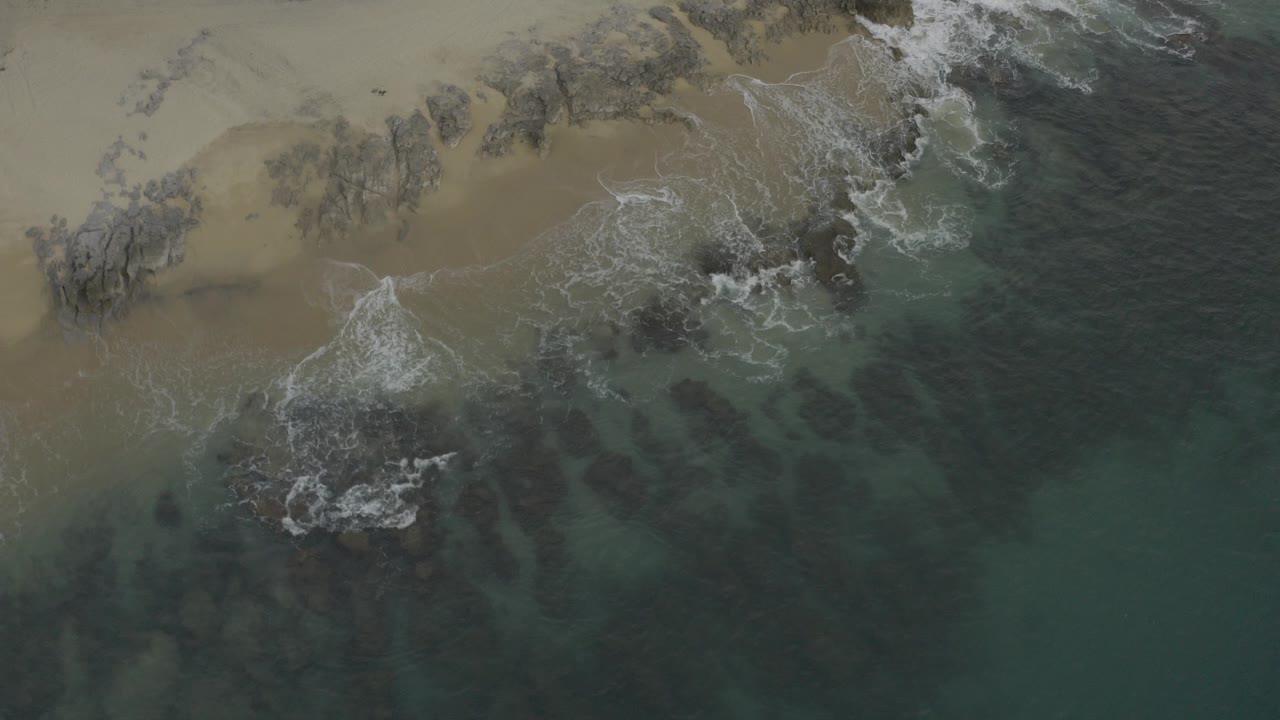 Overhead Baja California Sur beach frame with green water, white foam, tan sand, dark rocks, and no visible people at the start of the clip.