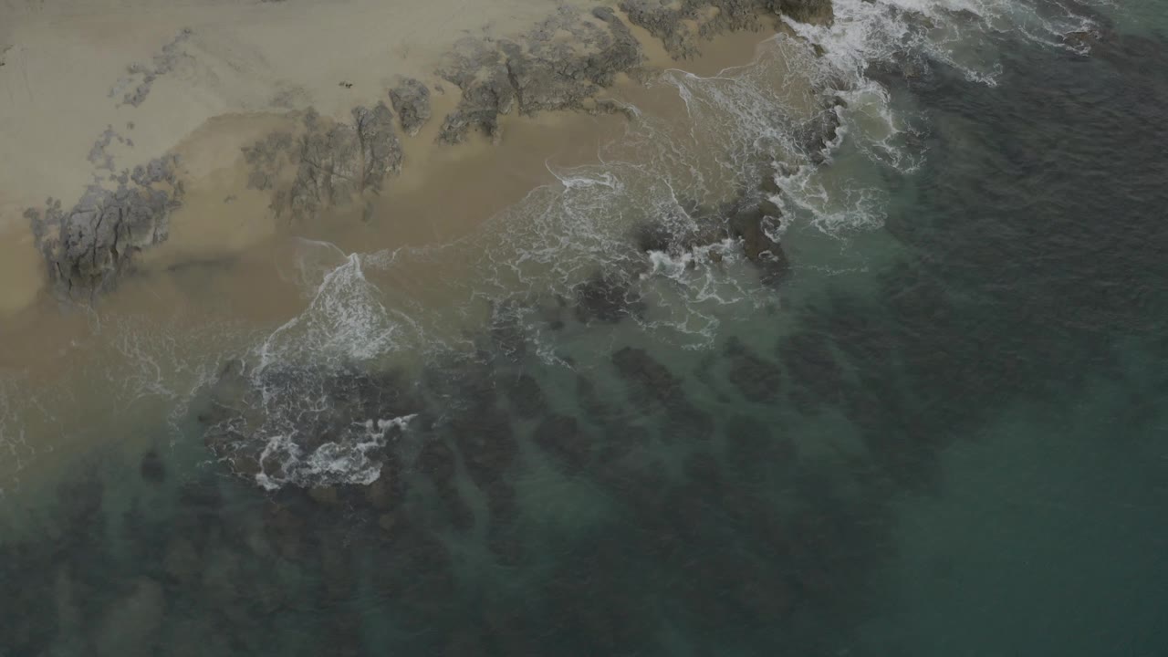 Aerial Baja California Sur shoreline view with foam spreading farther around a central rock and no visible people.