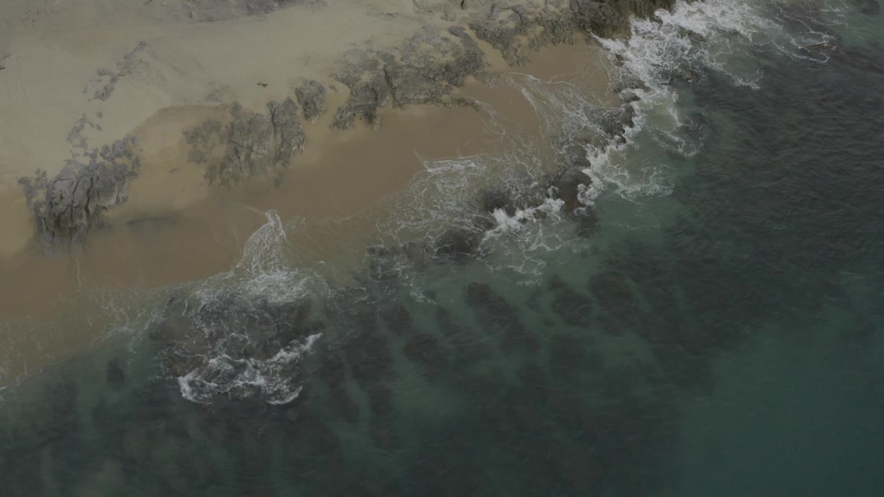 Overhead frame of Baja California Sur surf breaking against darker rocks near the sand, with green water below and no visible people.
