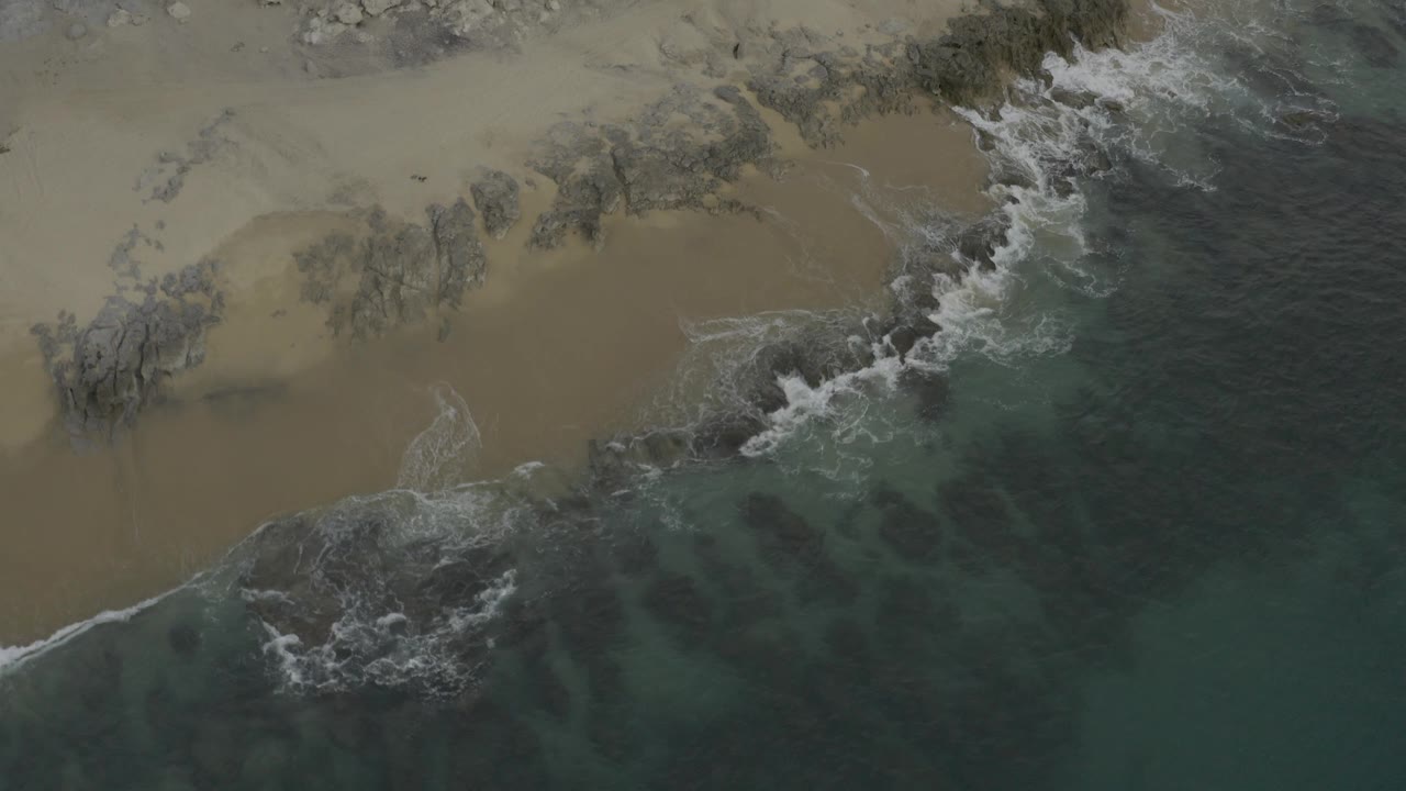 Baja California Sur beach seen from above, with tan sand across the upper frame, dark rocks near shore, green water below, and no visible people.