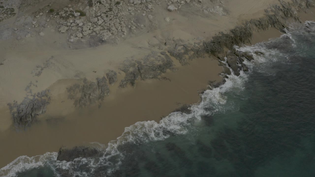 Overhead view of Baja California Sur shoreline where white surf curls across tan sand and dark rocks, with no visible people.