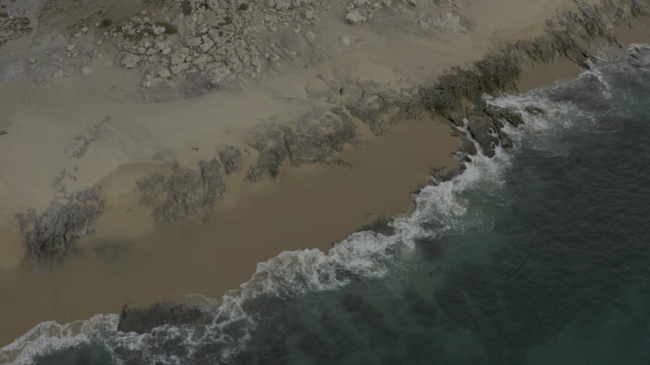 Aerial frame over Baja California Sur coast with a broad foam line spreading diagonally between teal water and tan sand, no visible people.