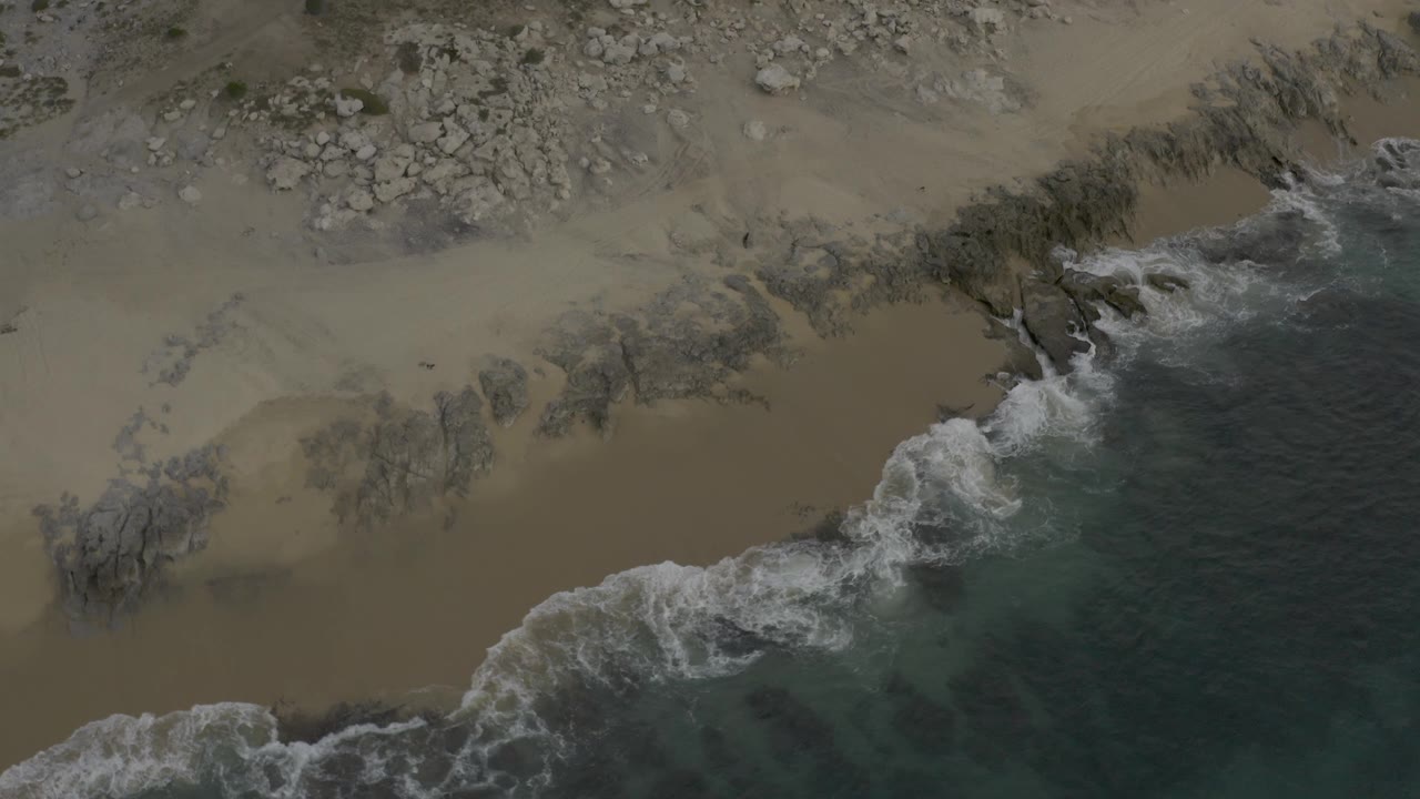 Top-down Baja California Sur beach view with wave wash wrapping around dark rock shelves and pale sand above, with no visible people.