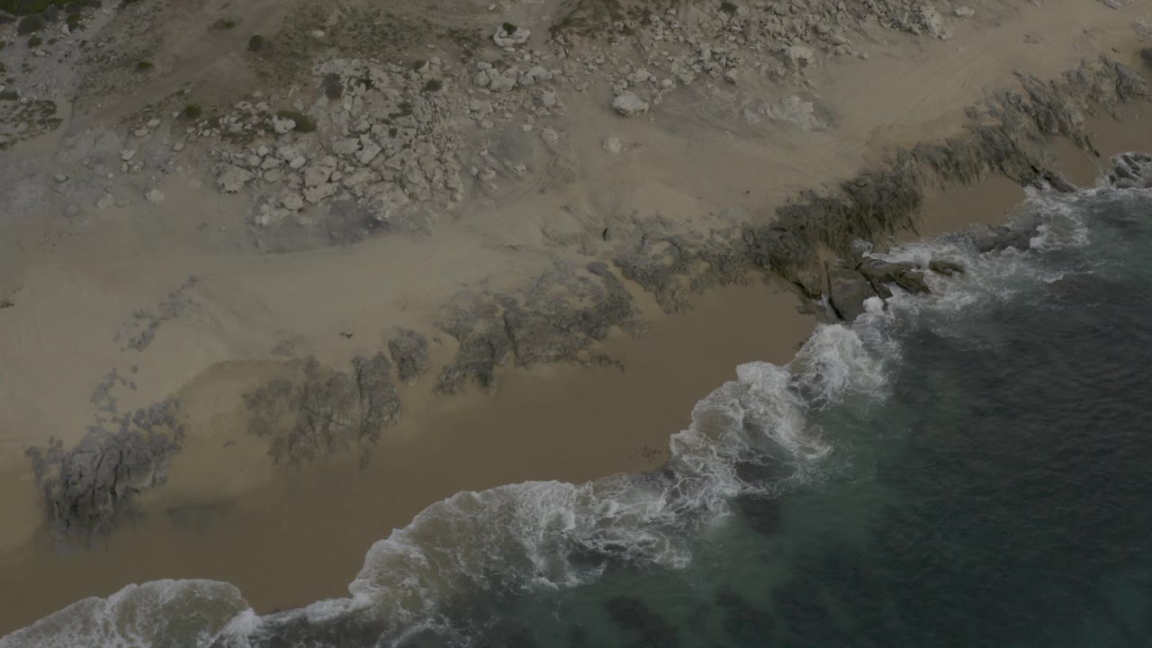 Aerial view of the Baja California Sur shoreline as whitewater reaches farther across the sandy edge below scattered rocks, no visible people.