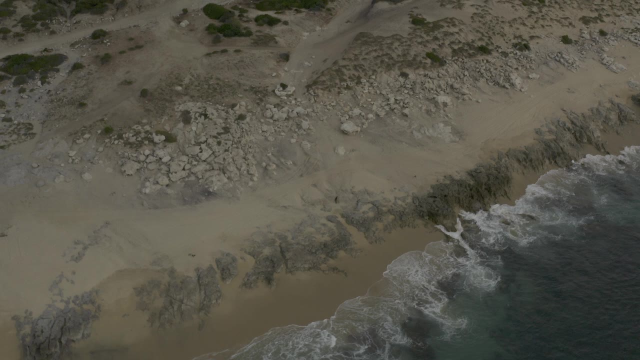 Overhead aerial frame of a Baja California Sur Mexico shoreline with tan sand, pale rocks, sparse green plants, and white surf entering from the lower right, with no visible people.