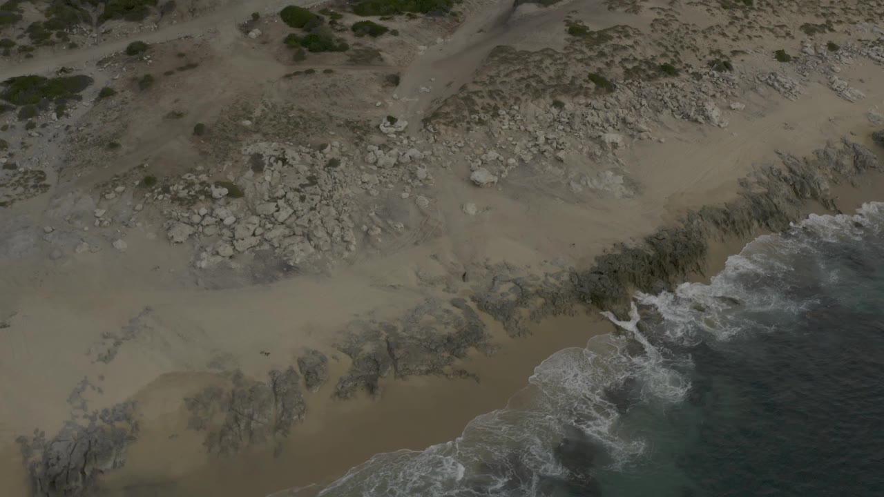 Aerial view over Baja California Sur Mexico coast showing a broad sandy area, scattered rock fields, low green vegetation near the top edge, and foamy waves along the right side, with no visible people.