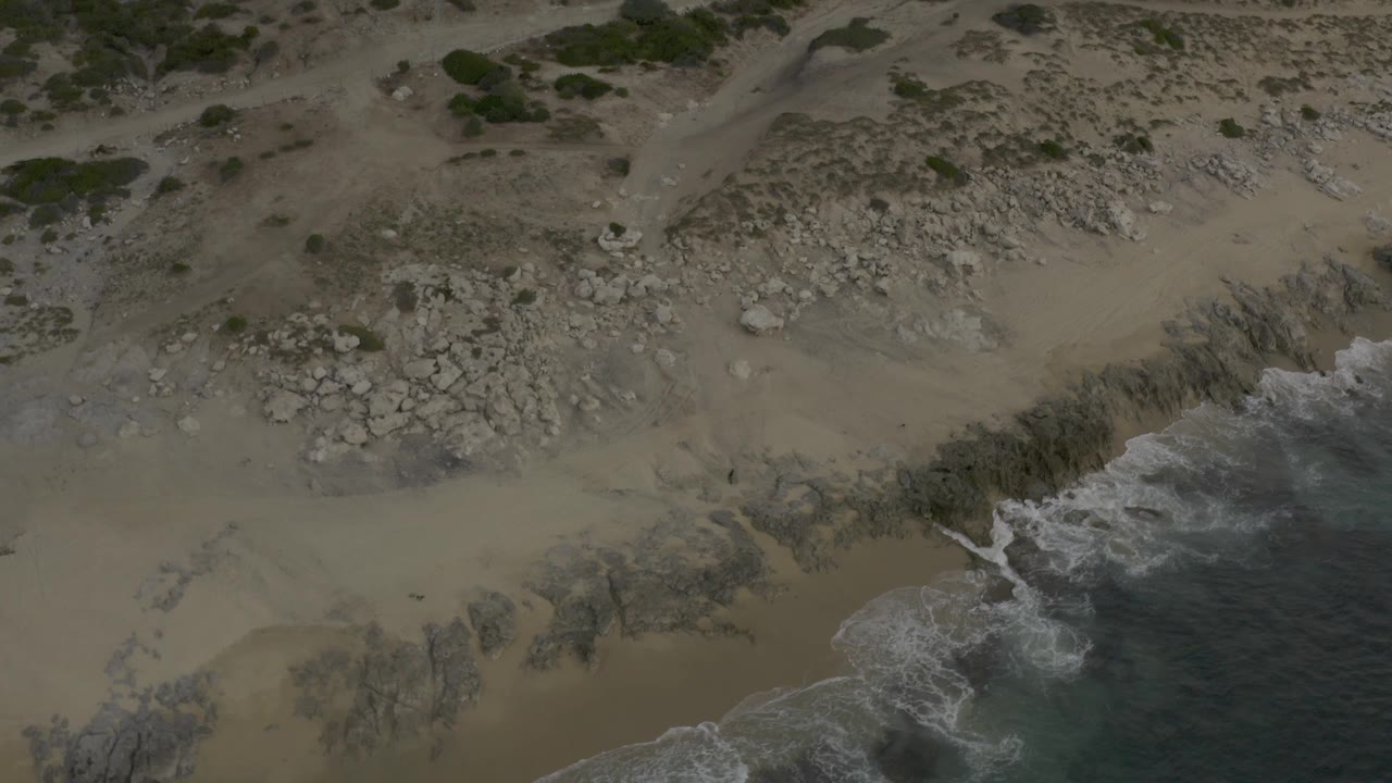 High overhead frame of the Baja California Sur Mexico seashore where darker water pushes white foam against jagged rocks beside tan sand and scrub patches, with no visible people.