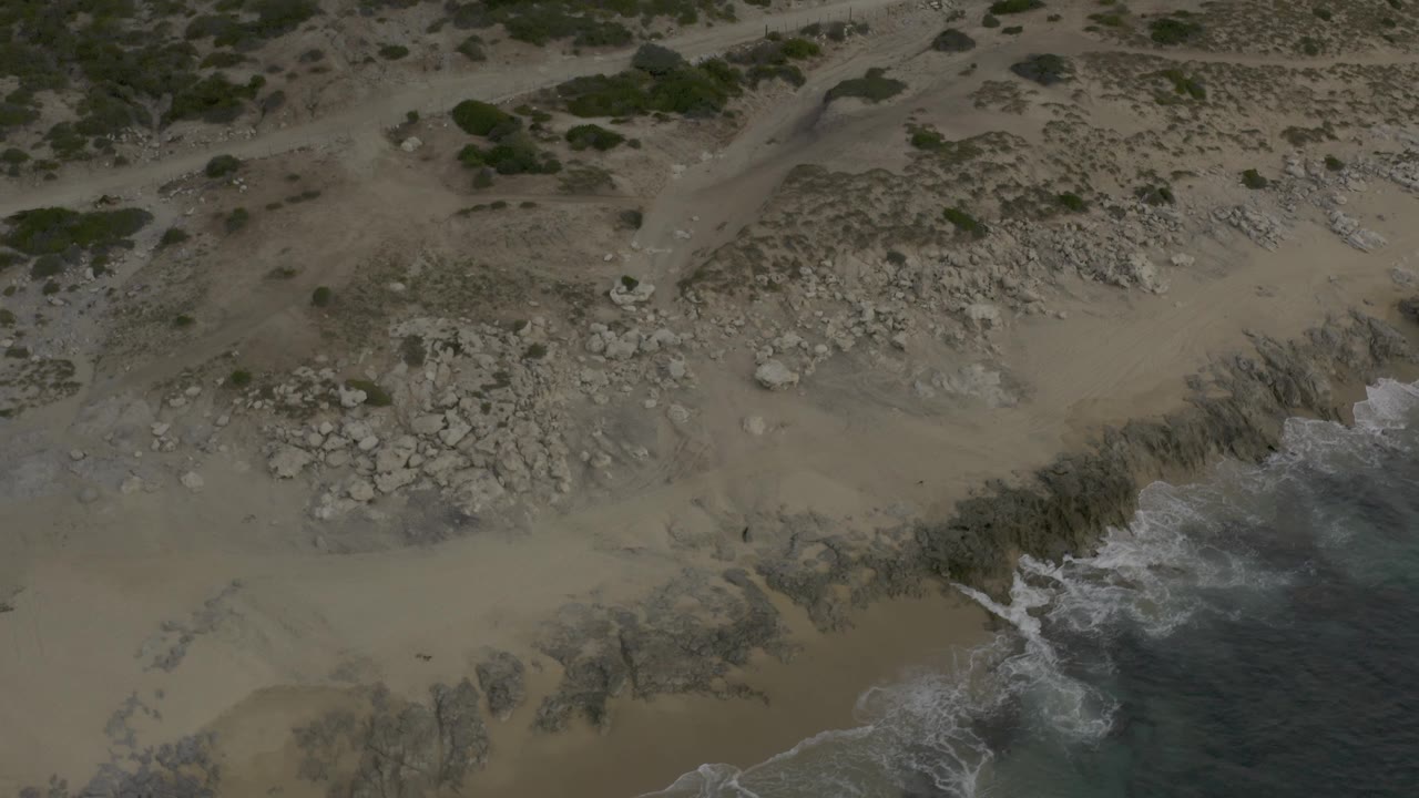 Drone-height view of Baja California Sur Mexico shoreline with a sandy upper beach, a central field of pale stones, and surf curling around the lower right rock shelf, with no visible people.