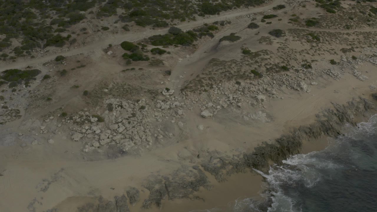 Aerial frame over Baja California Sur sand and scattered rocks, with green bushes above the beach and a small band of surf at the lower-right edge, no visible people.