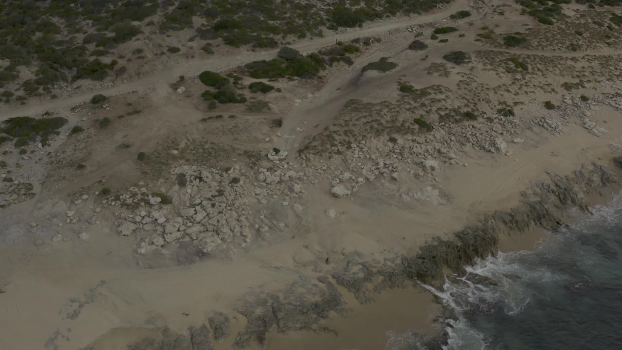 A Baja California Sur shoreline frame showing a broad patch of sand and stones, low bushes and a dirt track near the top, and white water curling along the right edge, no visible people.