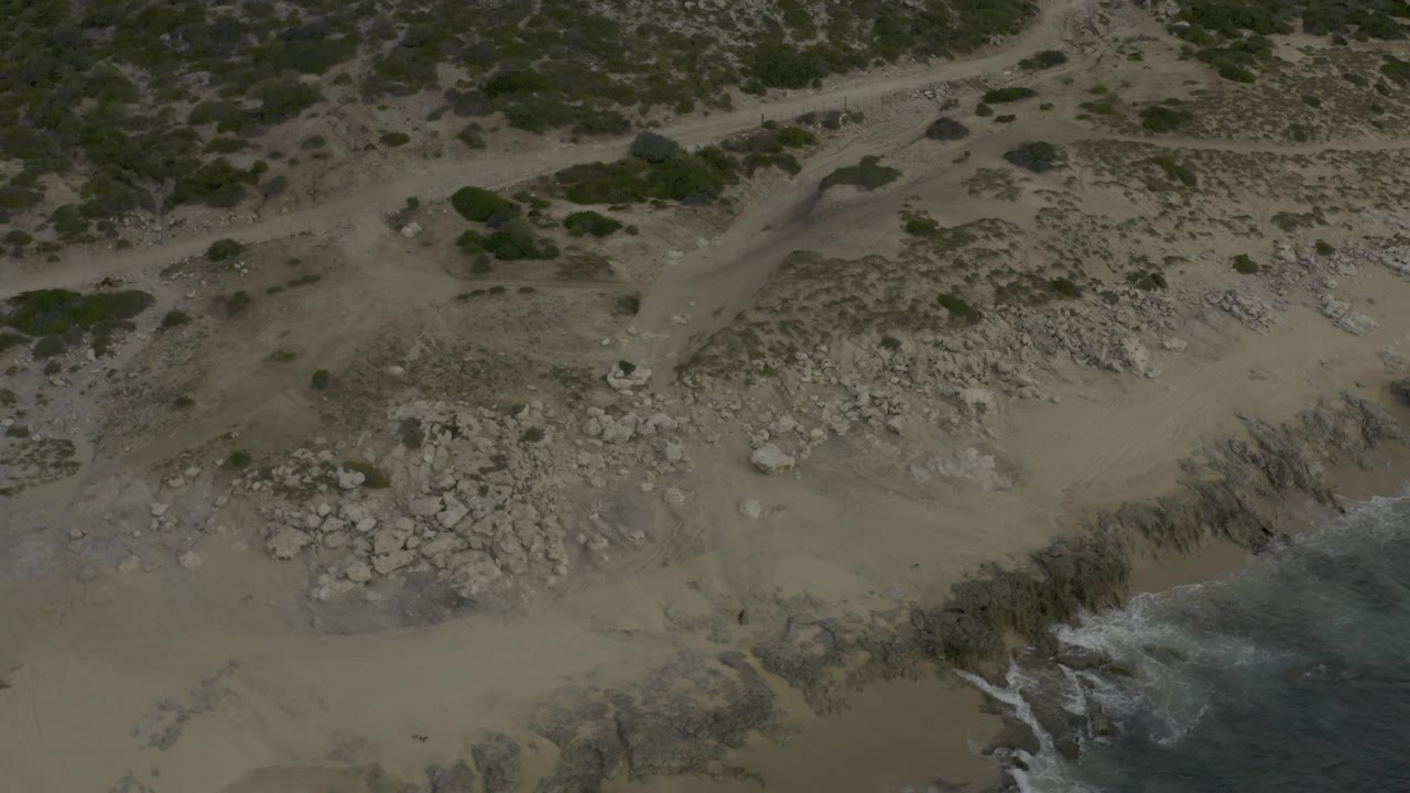 Overhead Baja California Sur beach view with the dirt track and scrub prominent at the top, broken rock across the center, and surf entering from the bottom-right corner, no visible people.