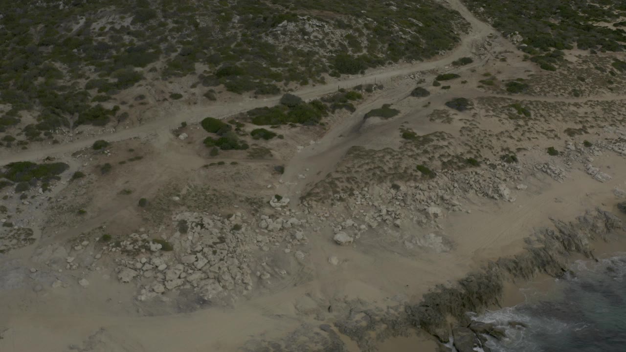 Aerial view of tan beach, grey rocks, scrubland, and a dirt track near the water