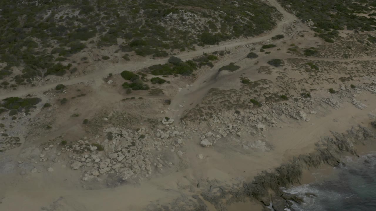 Aerial view in Baja California Sur with a central dirt path above a tan beach, scattered grey rocks, scrub vegetation, and no visible people.