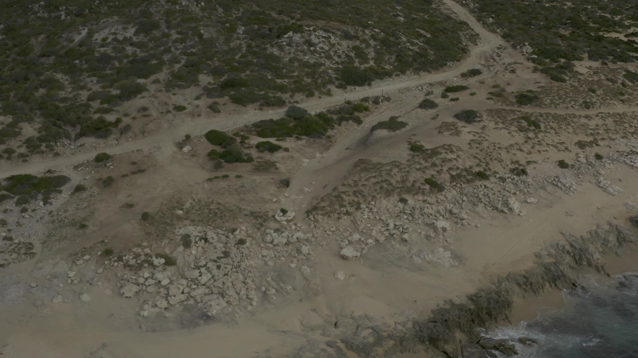 Baja California Sur beach frame with scrubland across the top, pale rocks and tan sand below, and a narrow blue-green water edge, with no visible people.