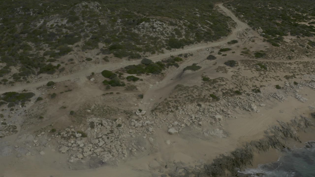 Aerial frame from Baja California Sur showing crossing dirt tracks, tan beach, grey boulders, scrub-covered ground, and no visible people.
